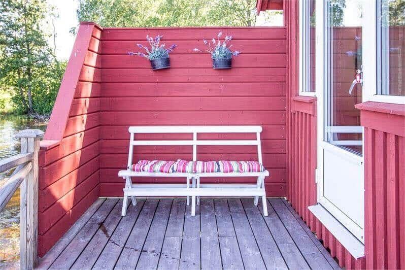 Roter Holzveranda mit weißem Stuhl und Kissen, zwei Blumenkästen an der Wand.