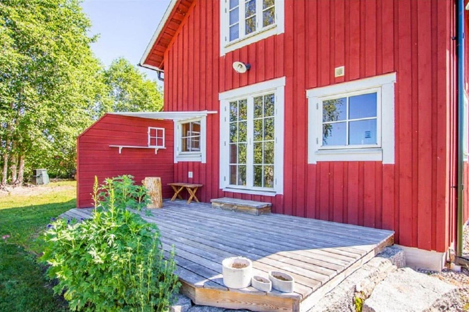 Rotes Holzhaus mit Terrasse, Fenstern und Garten. Holzbank und Pflanzen sind sichtbar.