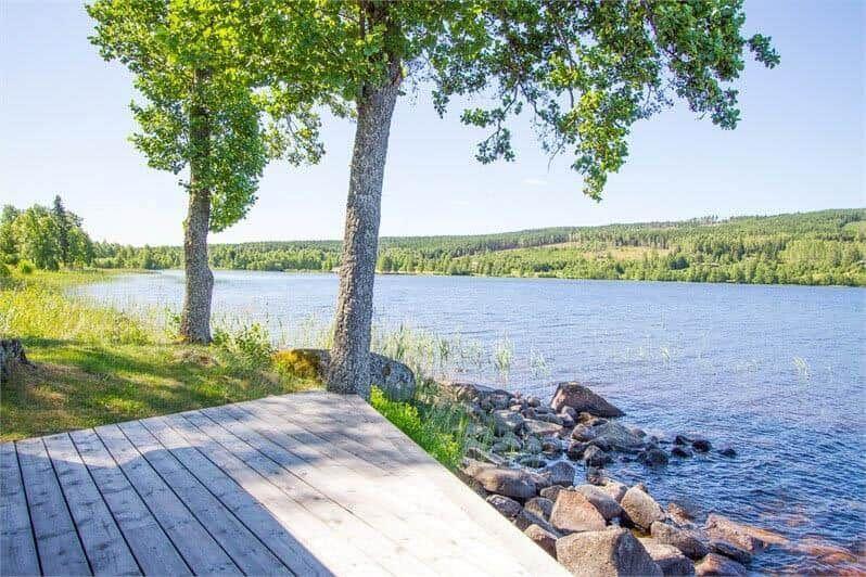 Holzterrasse am See mit Bäumen und Steinufer.