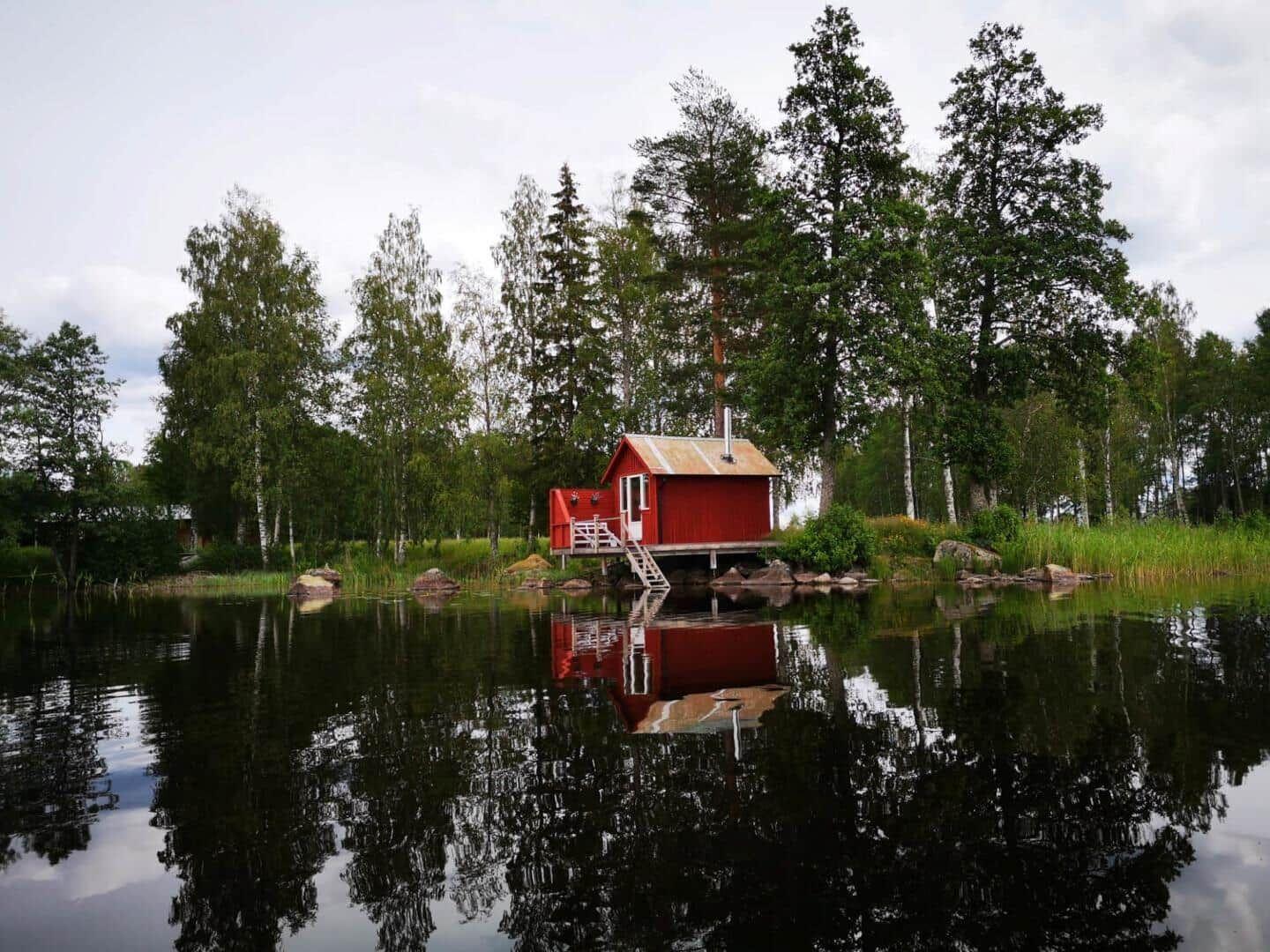 Rotes Holzhaus am See mit Reflexion und umgebenden Bäumen.