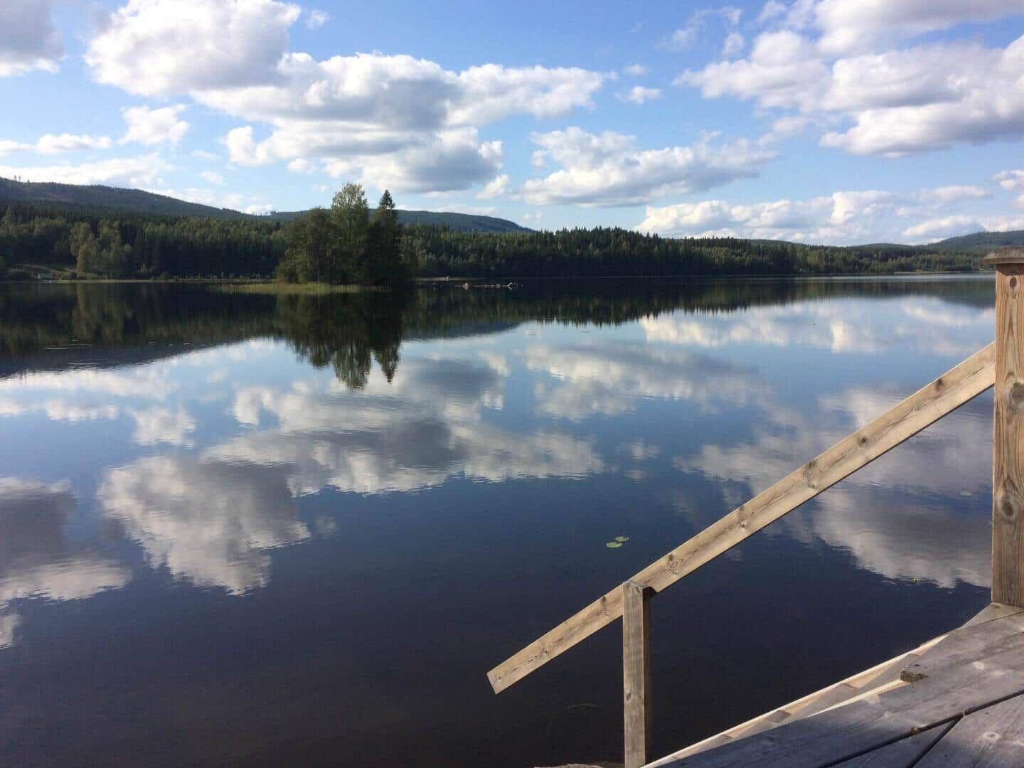Ein Holzsteg führt zu einem ruhigen See mit Spiegelung von Himmel und Bäumen.