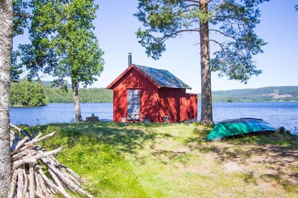 Rotes Holzhaus am See mit Boot und Feuerholz unter Bäumen