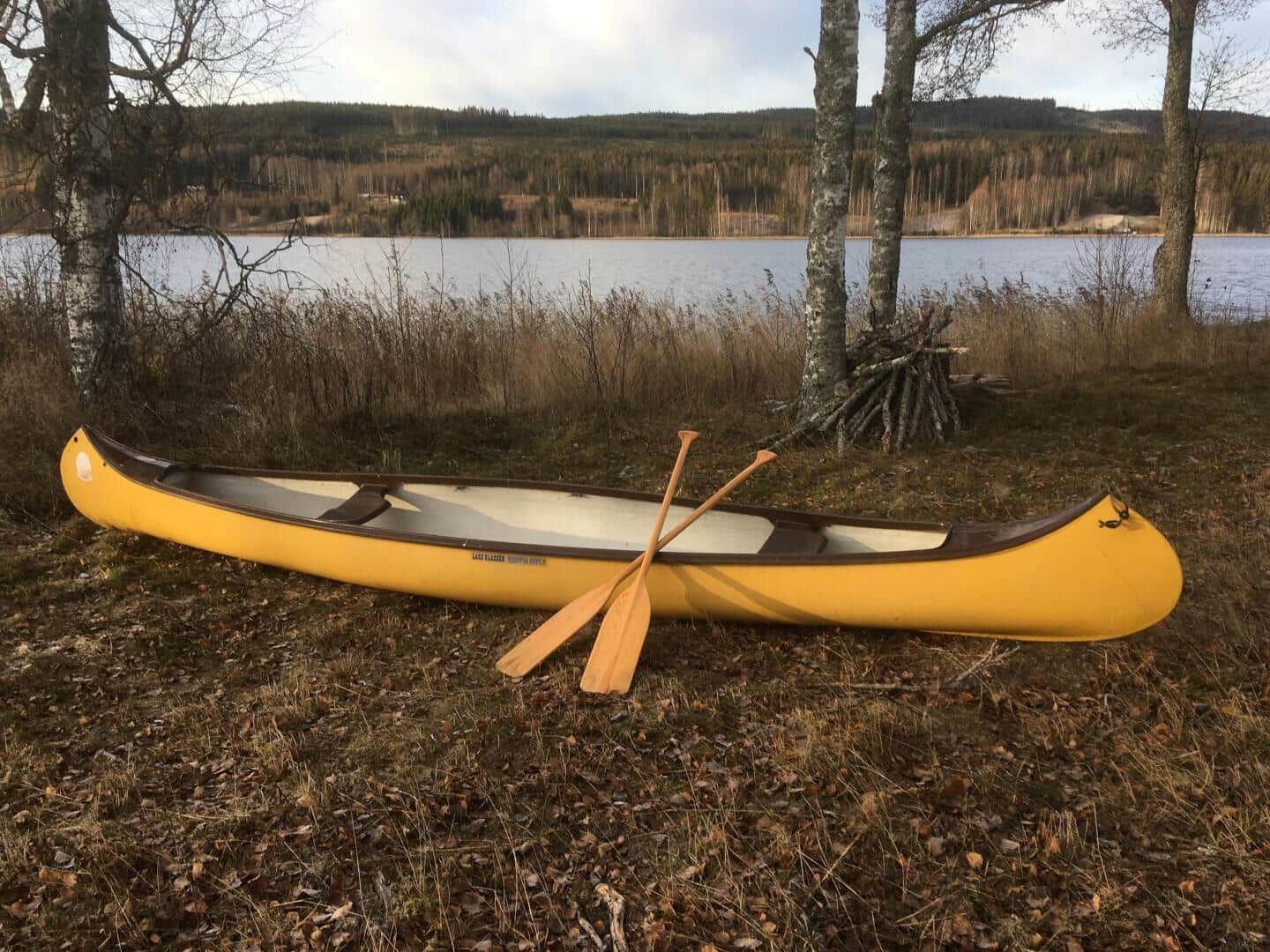 Ein gelbes Kanu mit zwei Ruder liegt am Ufer eines Sees. Im Hintergrund sind Bäume und Wasser zu sehen.