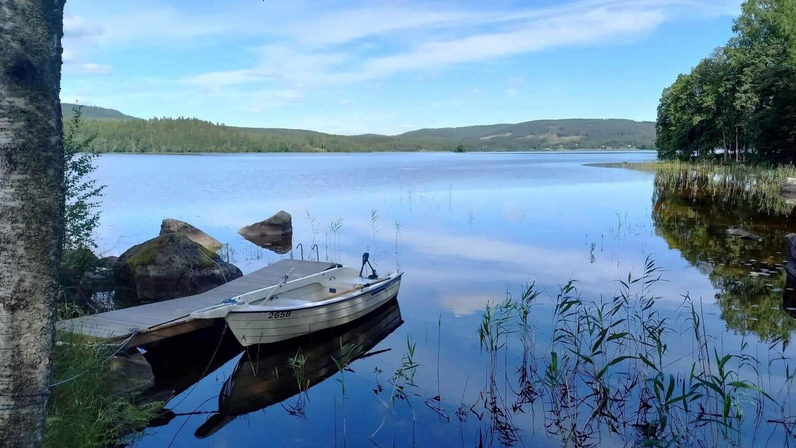 Ein kleines Boot liegt an einem Holzsteg am Ufer eines ruhigen Sees mit Bergen im Hintergrund.