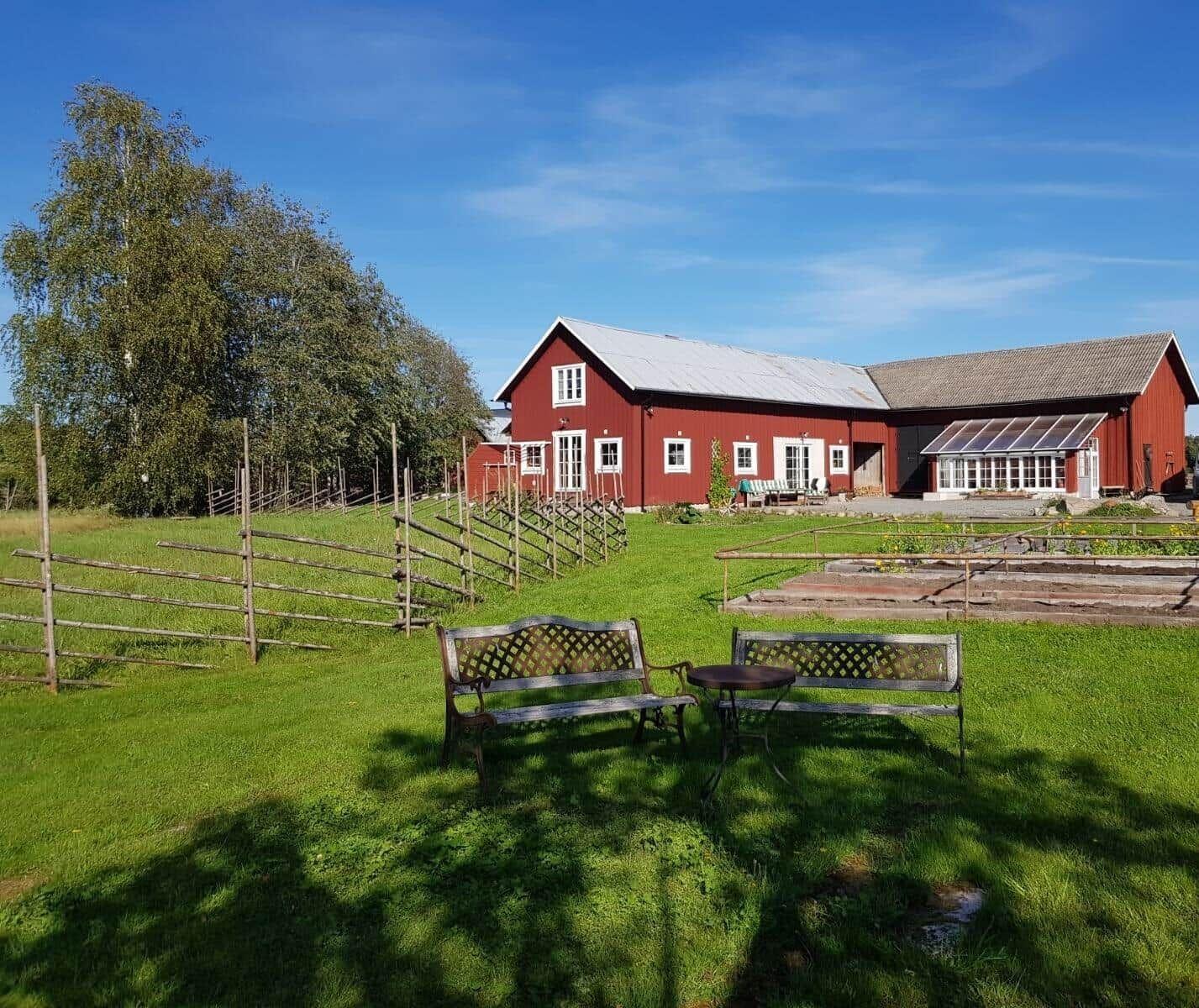 Roter Bauernhof mit Garten und Bänken unter blauem Himmel.