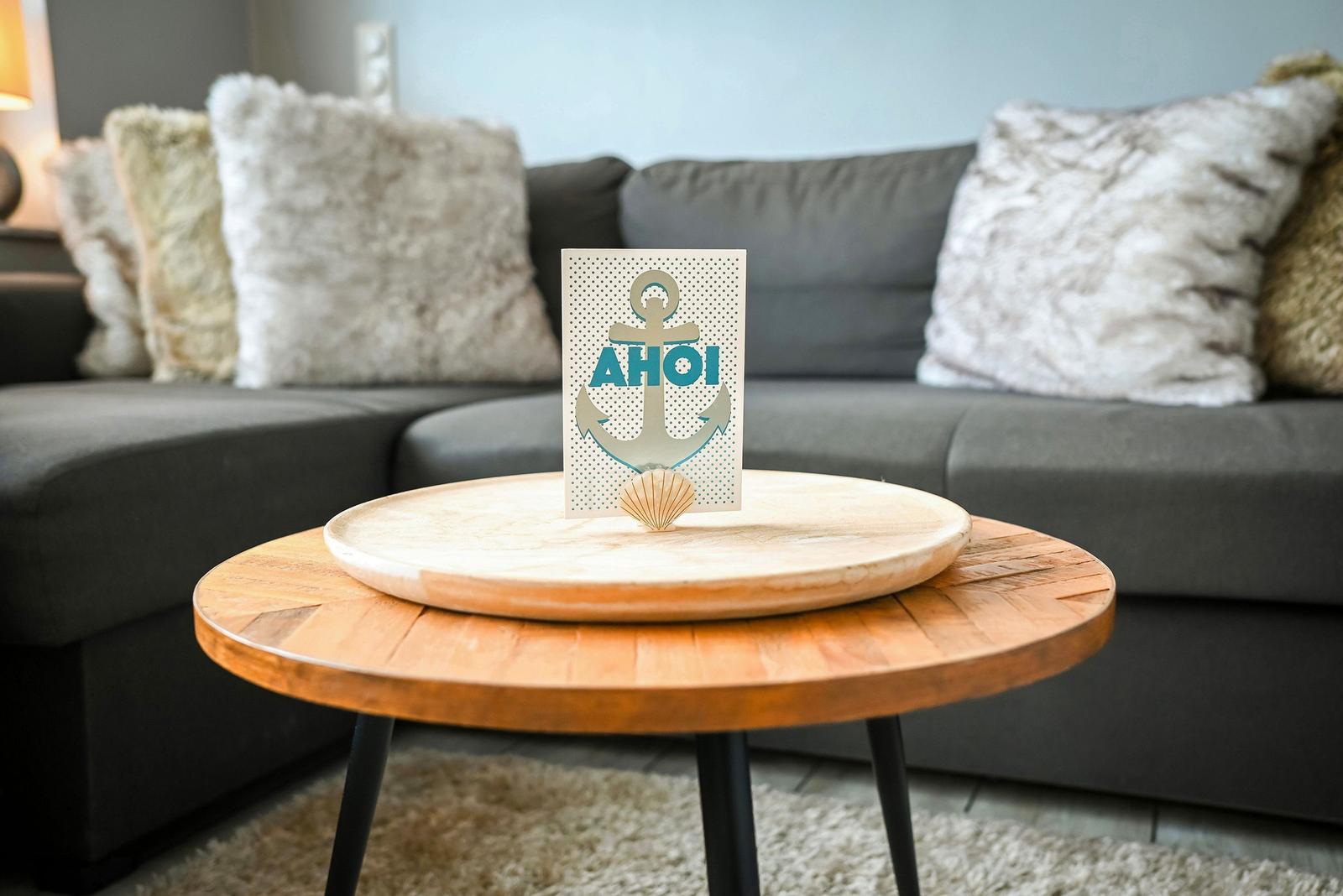Ein runder Holztisch mit einer Karte und einer Muschel. Hintergrund: Sofa mit Kissen.