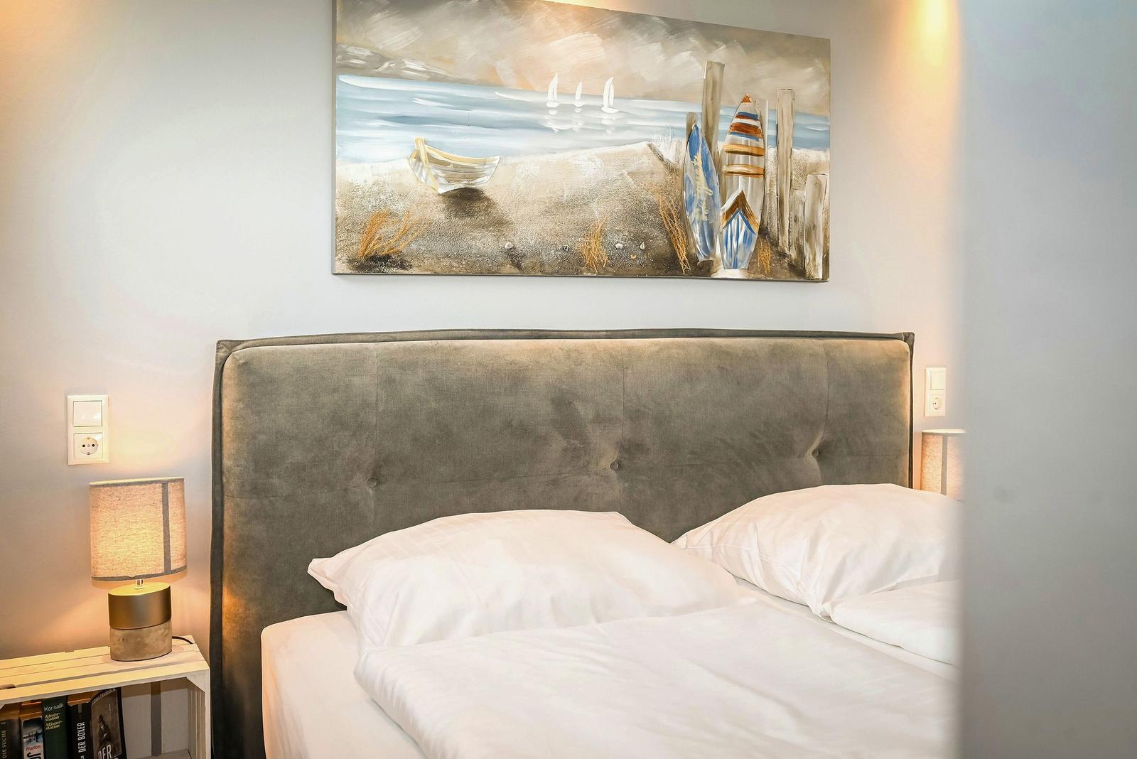 Ein Bett mit weißer Bettwäsche und einem grauen Kopfteil. Über dem Bett hängt ein Strandgemälde.