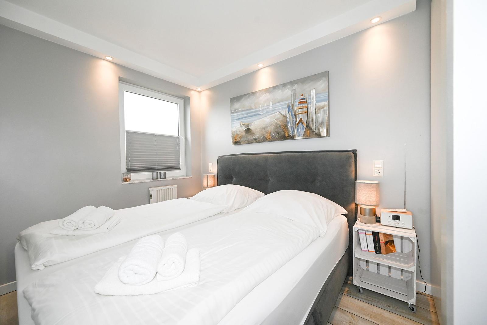 Schlafzimmer mit Bett, Nachttisch und Wandbild. Fenster mit Jalousie.