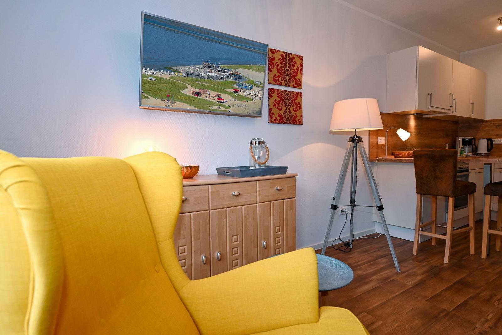 Ein Raum mit gelbem Sessel, Küchenbereich und Wandbild.