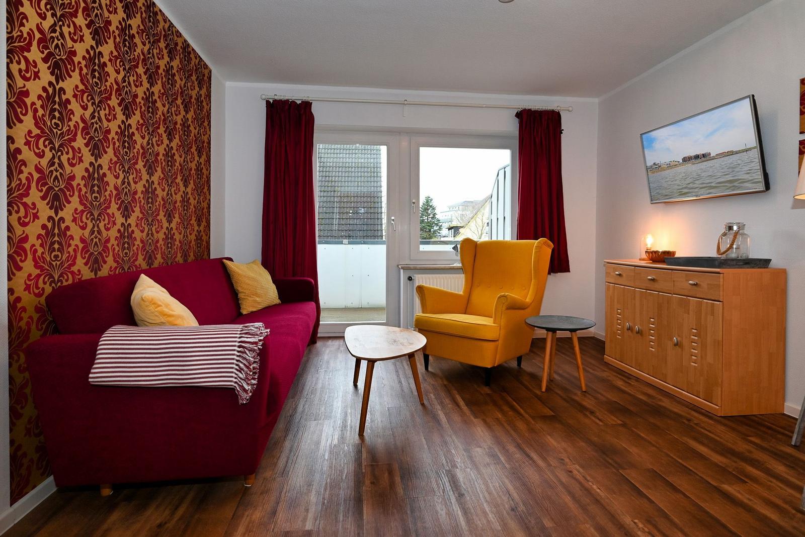 Ein Wohnzimmer mit rotem Sofa, gelber Sessel und Holzboden. An der Wand hängt ein Fernseher.