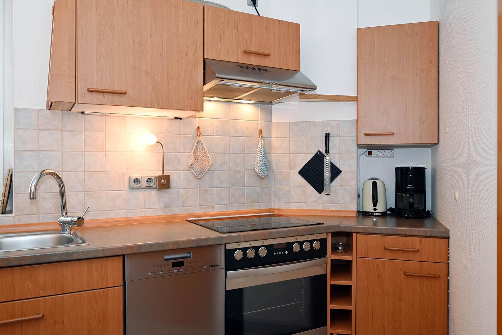 Küche mit Holzschränken, Spüle, Kochfeld und Backofen. Auf Arbeitsfläche stehen Kaffeemaschine und Wasserkocher.