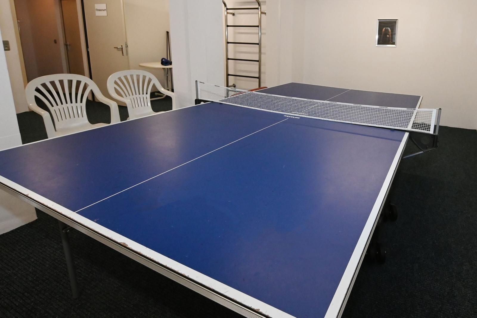 Tischtennis-Tisch mit zwei weißen Stühlen in einem Raum mit dunklem Teppichboden.