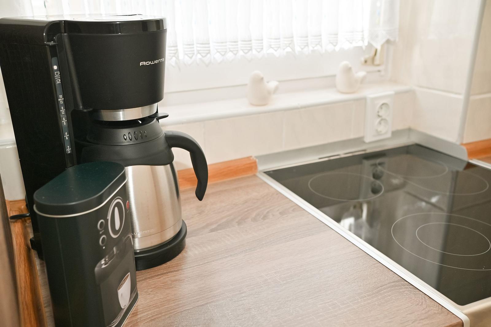 Küchenzeile mit Kaffeemaschine, Kaffeekanne und Herd. Fenster mit weißen Vorhängen.