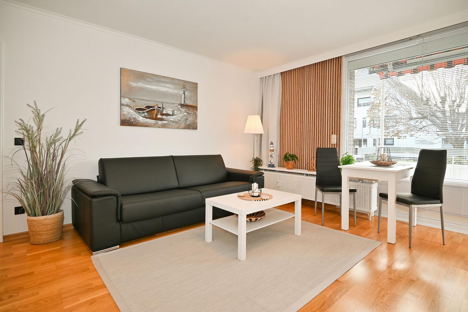 Wohn- und Essbereich mit Sofa, Tisch und Stühlen. Holzboden und Fenster mit Jalousien.