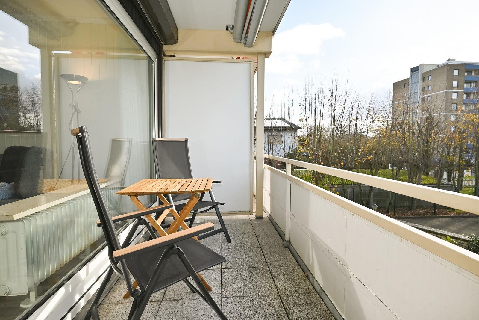 Terrasse mit Tisch und zwei Stühlen, Blick auf Bäume und Gebäude.