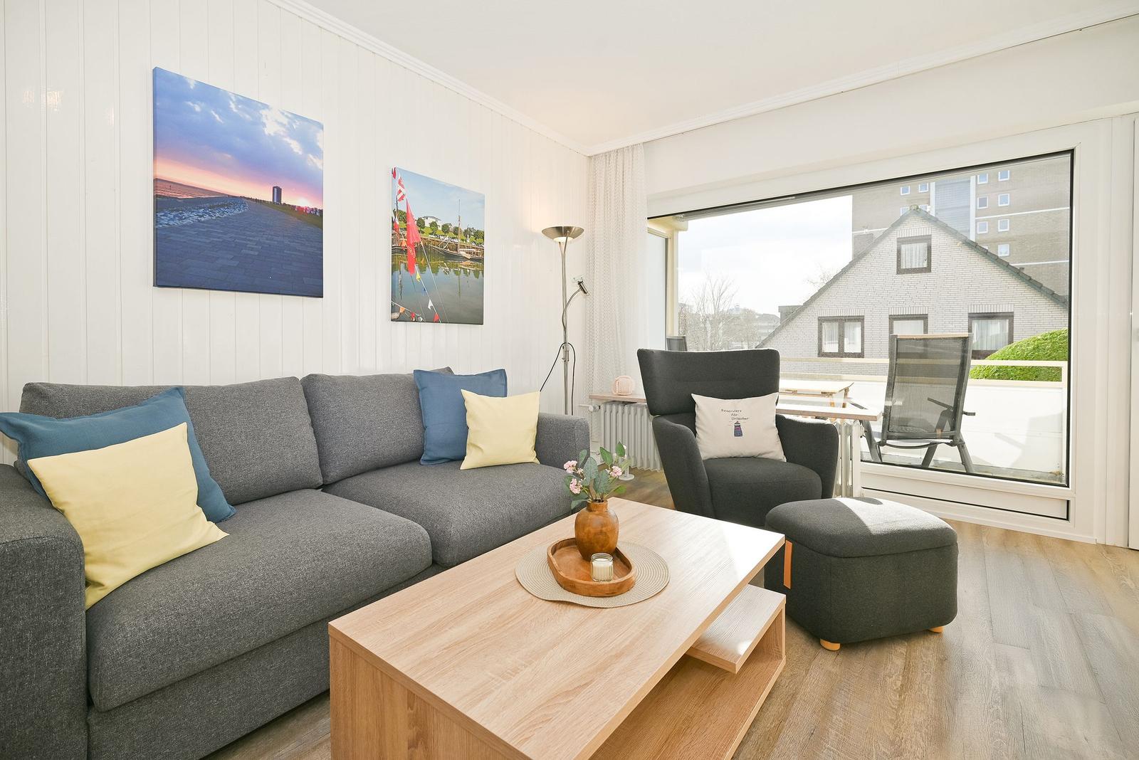 Wohnzimmer mit Sofa, Sessel und Tisch. Blick auf Balkon mit Stuhl. Wandbilder und Holzboden.