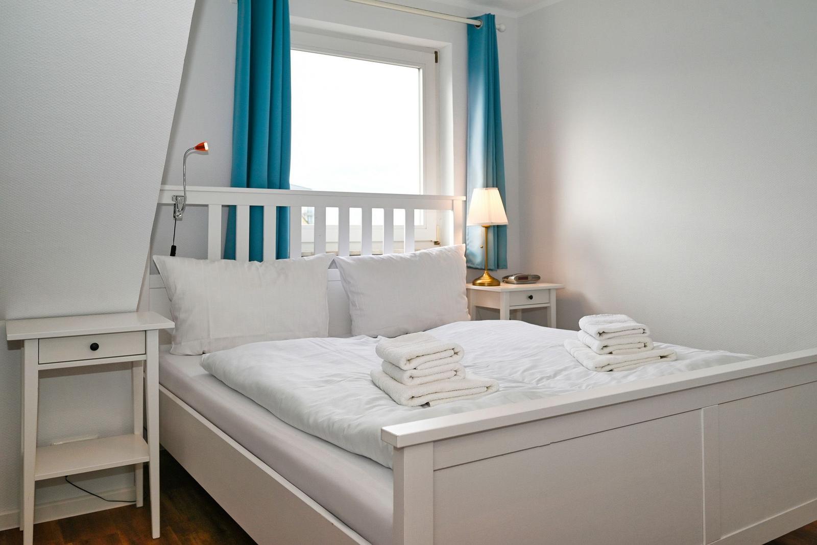 Schlafzimmer mit weißem Bett, zwei Nachttischen und blauen Vorhängen an einem Fenster.