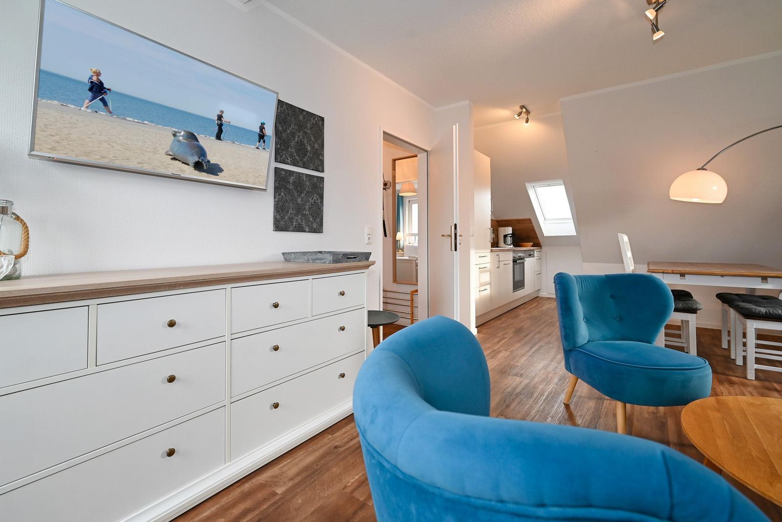 Wohnbereich mit blauen Sesseln, Küchenbereich und Fernseher an der Wand.