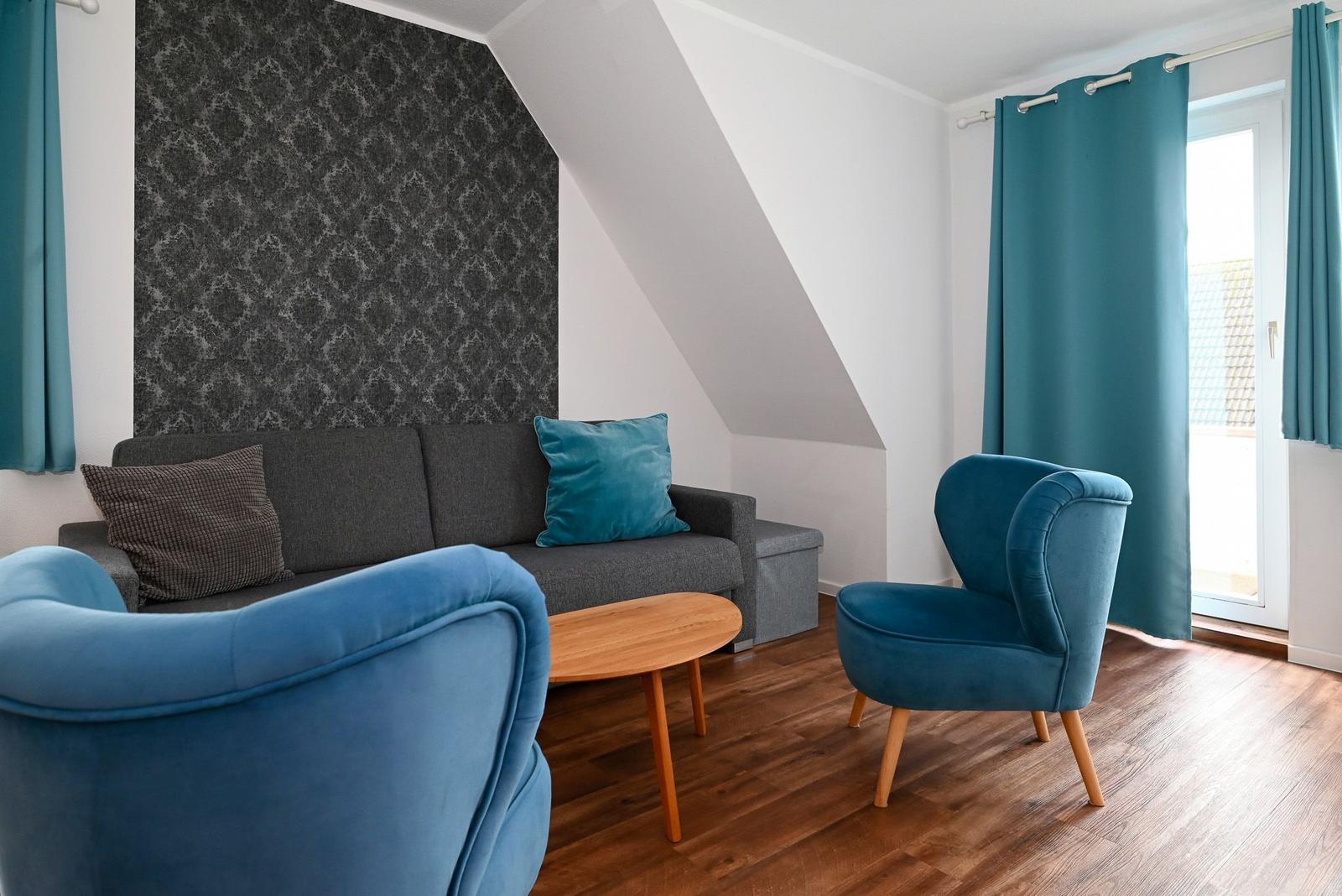 Wohnzimmer mit Sofa, zwei blauen Sesseln und Holztisch unter Dachschräge.