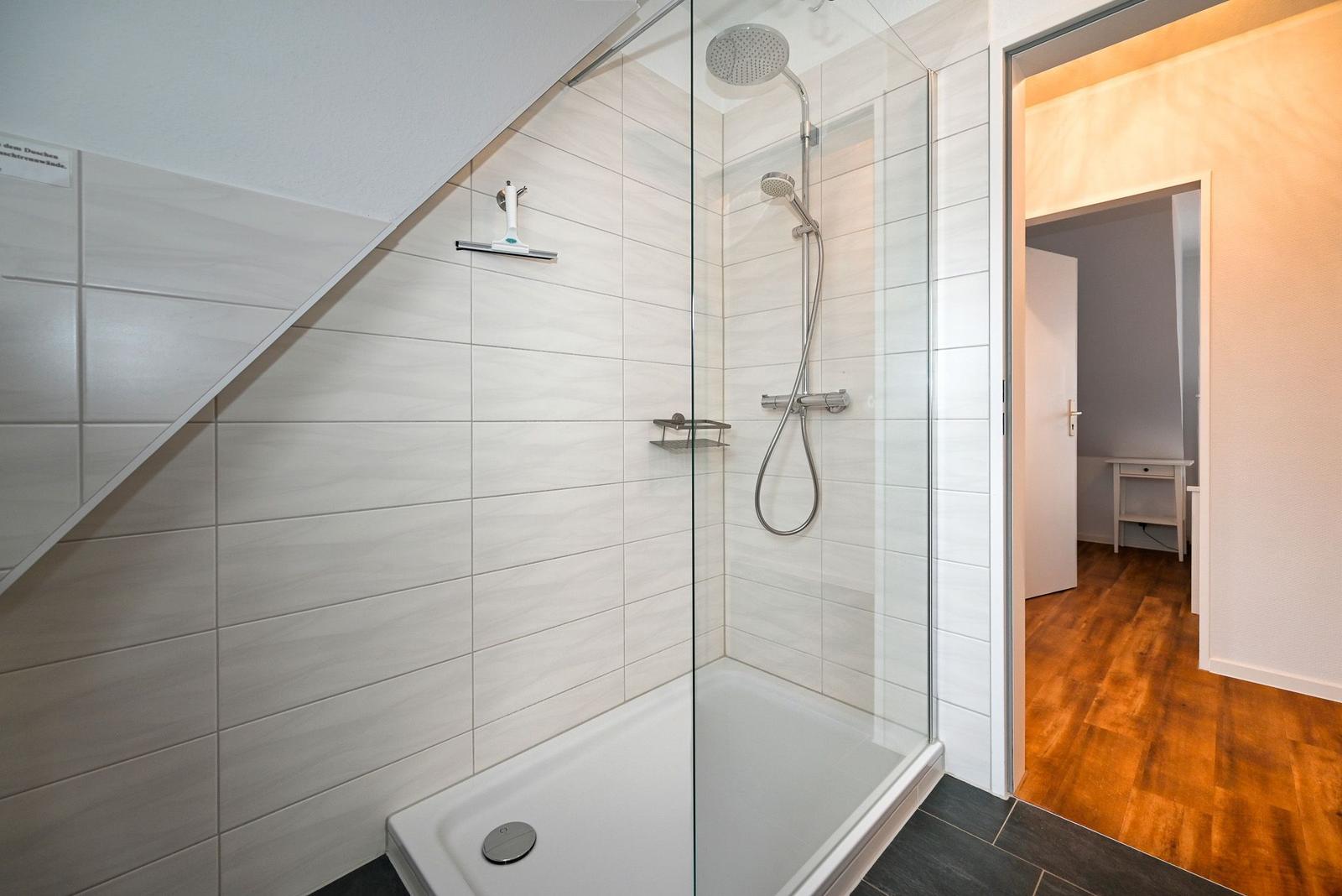Dusche mit Glaswand, Duschbrause und Handbrause. Tür führt in einen Raum mit Holzboden.