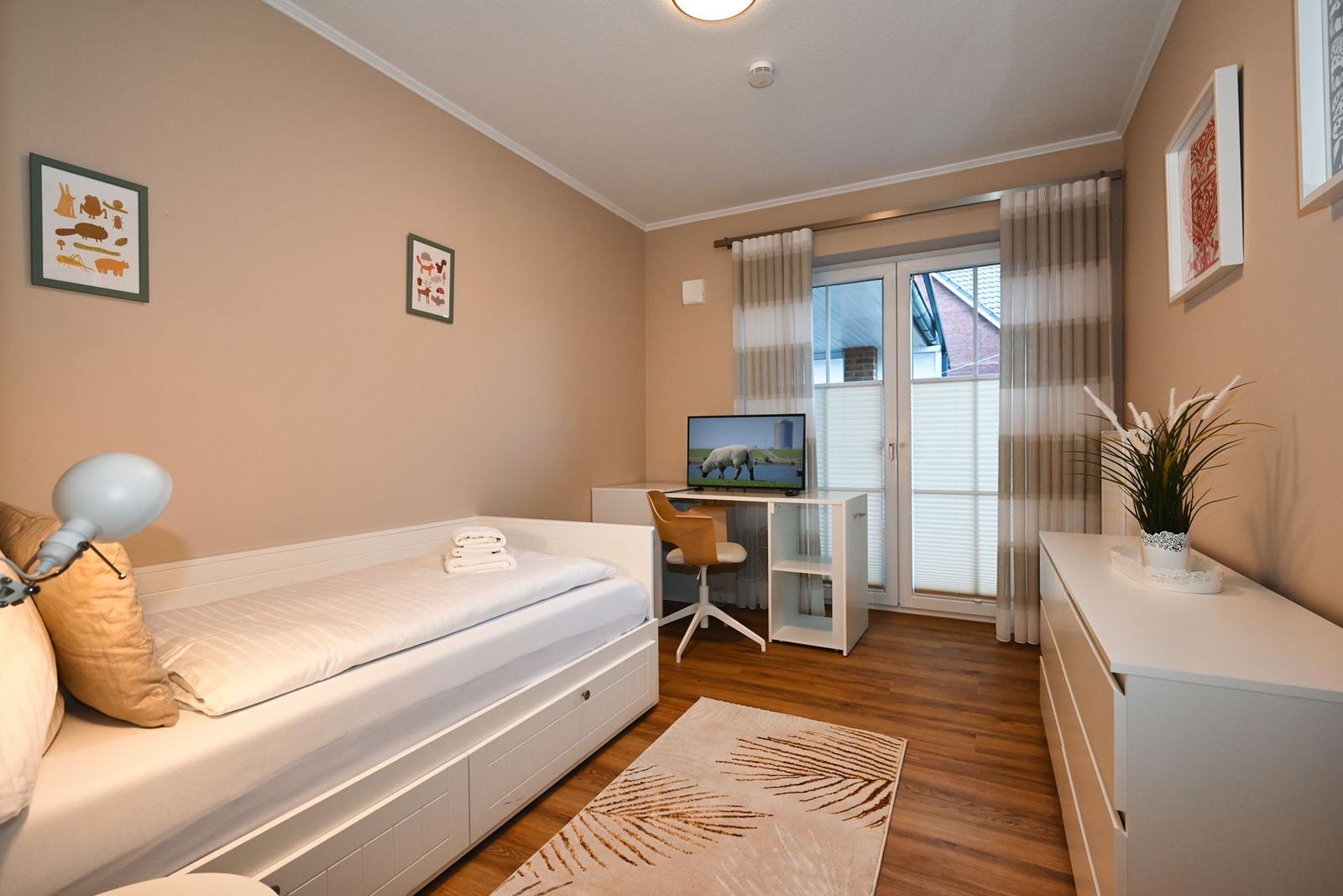 Schlafzimmer mit Bett, Schreibtisch und TV. Fenster mit Jalousien und Vorhängen.