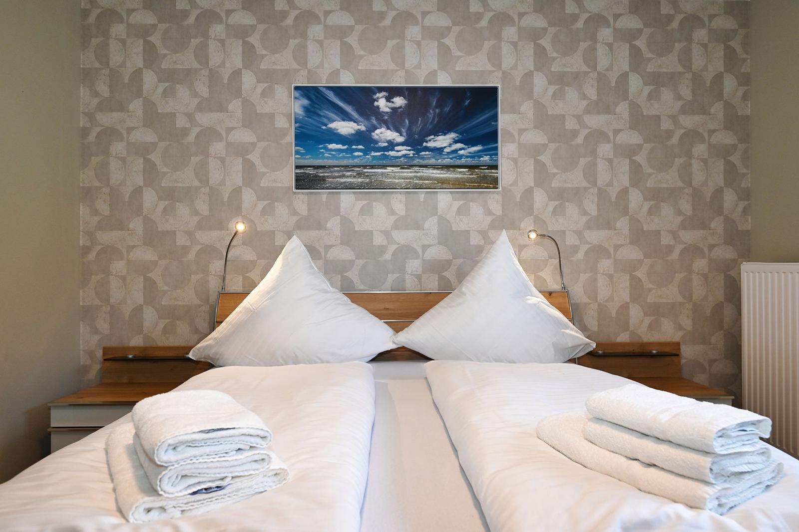 Doppelbett mit weißen Laken, zwei Kissen und gefalteten Handtüchern. Wandbild zeigt Strand und Himmel.