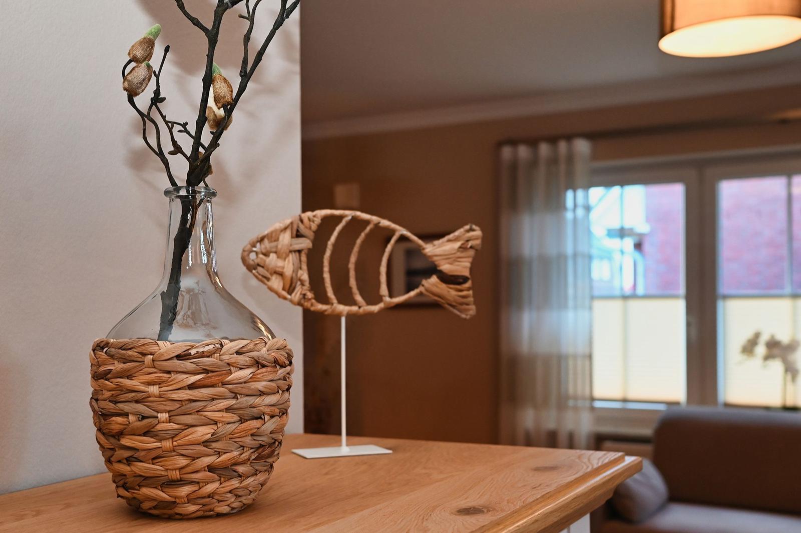 Auf dem Tisch steht eine geflochtene Vase mit Zweigen und eine Fischdekoration.