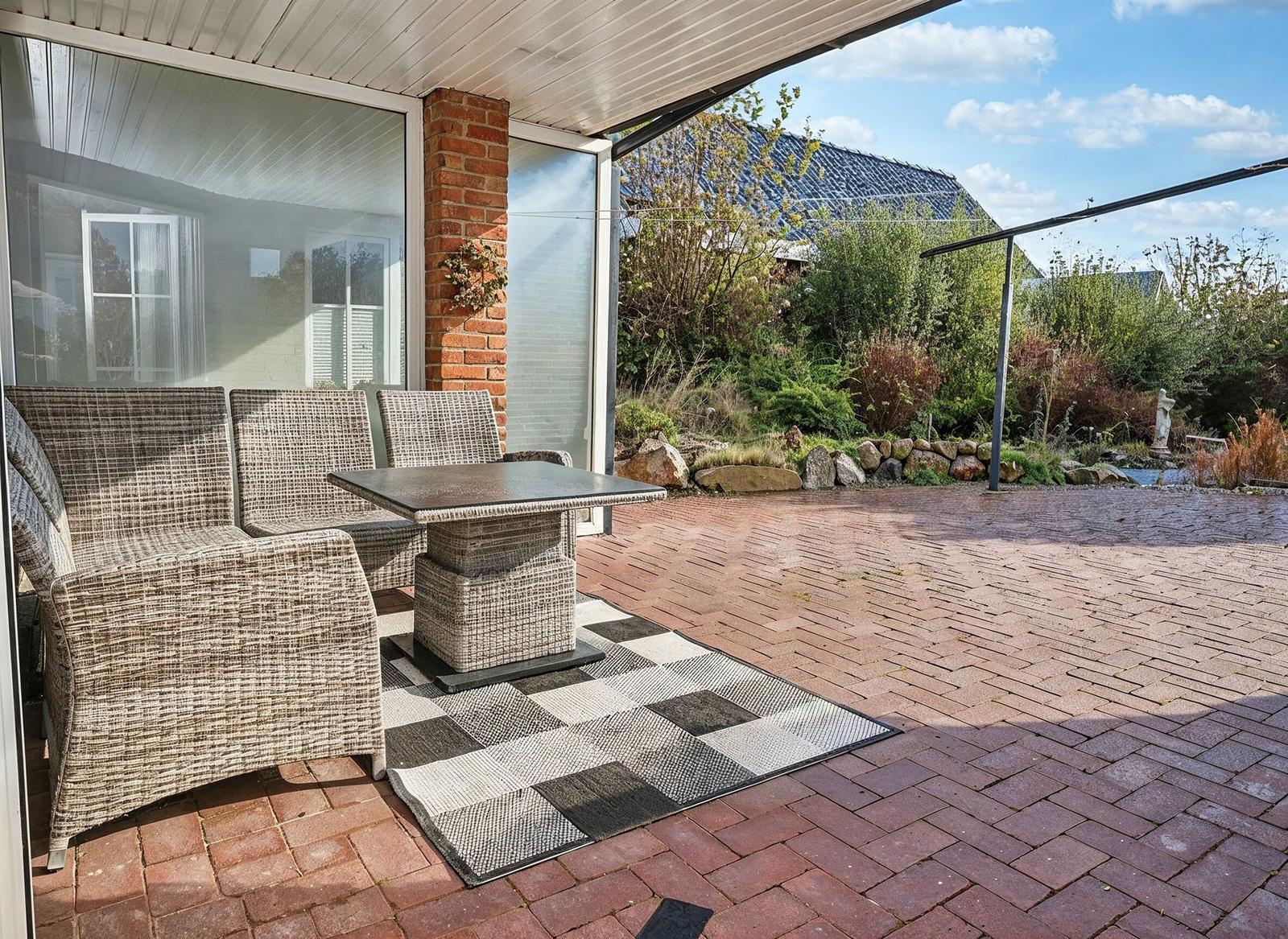 Terrasse mit Sitzgruppe, Tisch und Teppich auf Steinplatten. Hintergrund mit Garten und Steinmauer.