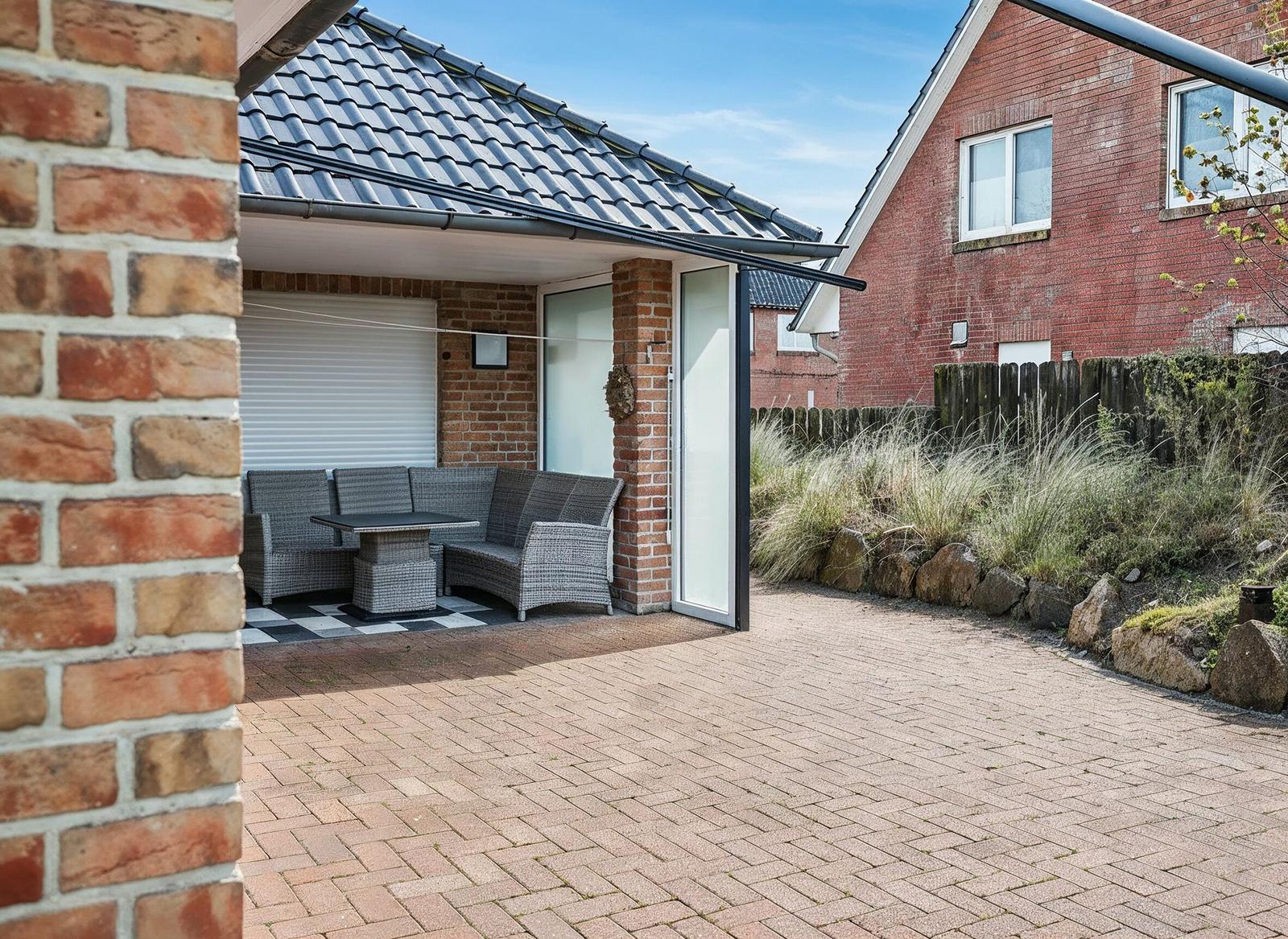 Terrasse mit Sitzgruppe unter Dach, gepflasterter Hof und Gartenbereich mit Steinen.