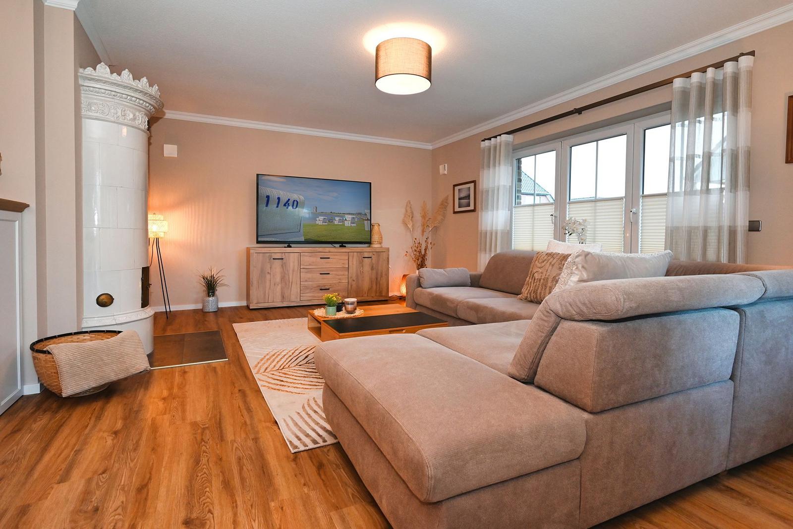 Wohnzimmer mit Sofa, Fernseher und Kaminofen. Holzboden und große Fenster.