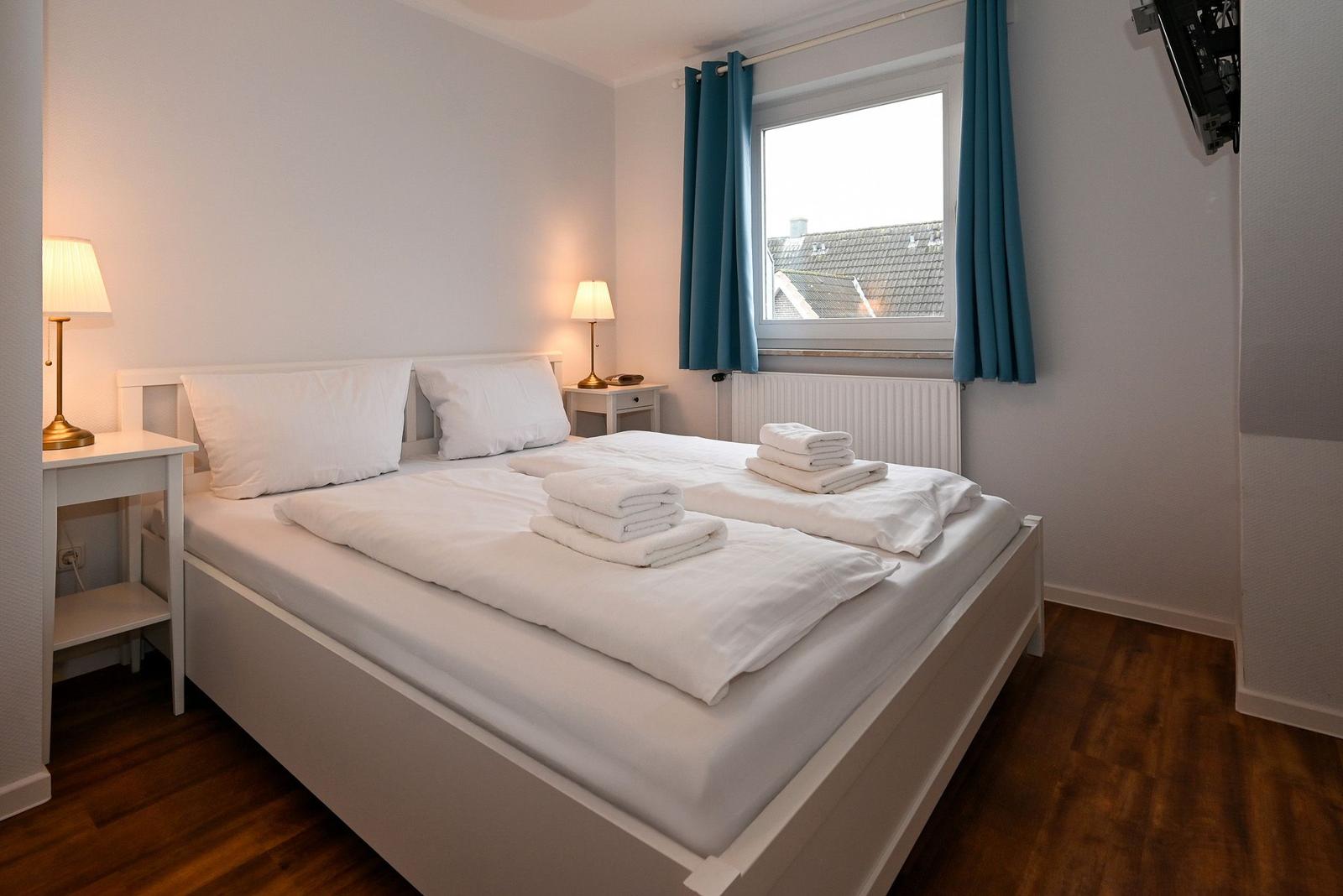 Schlafzimmer mit Doppelbett, zwei Nachttischen und Fenster mit blauen Vorhängen.