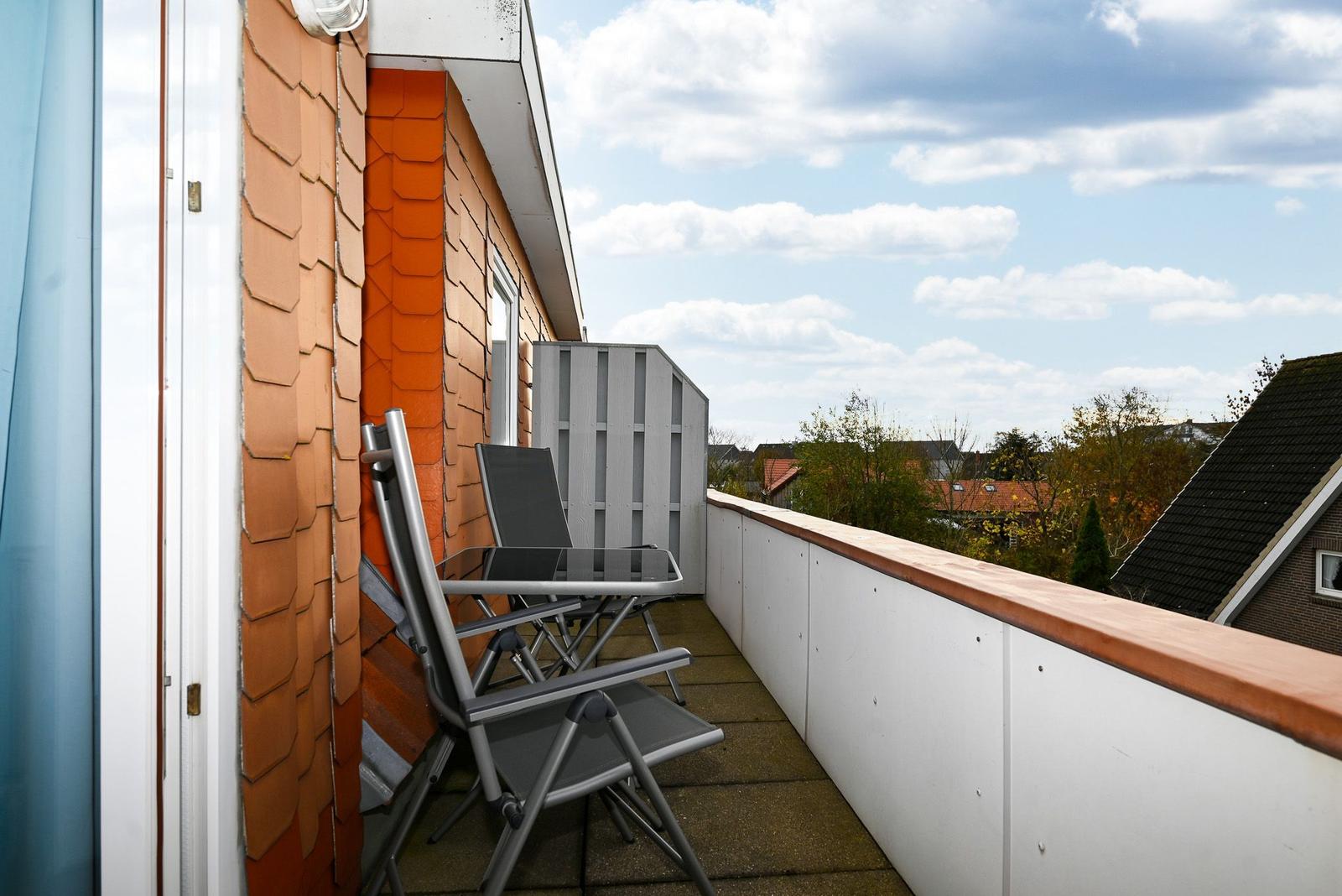 Terrasse mit Stuhl und Tisch, Blick auf Dächer und Bäume unter blauem Himmel.