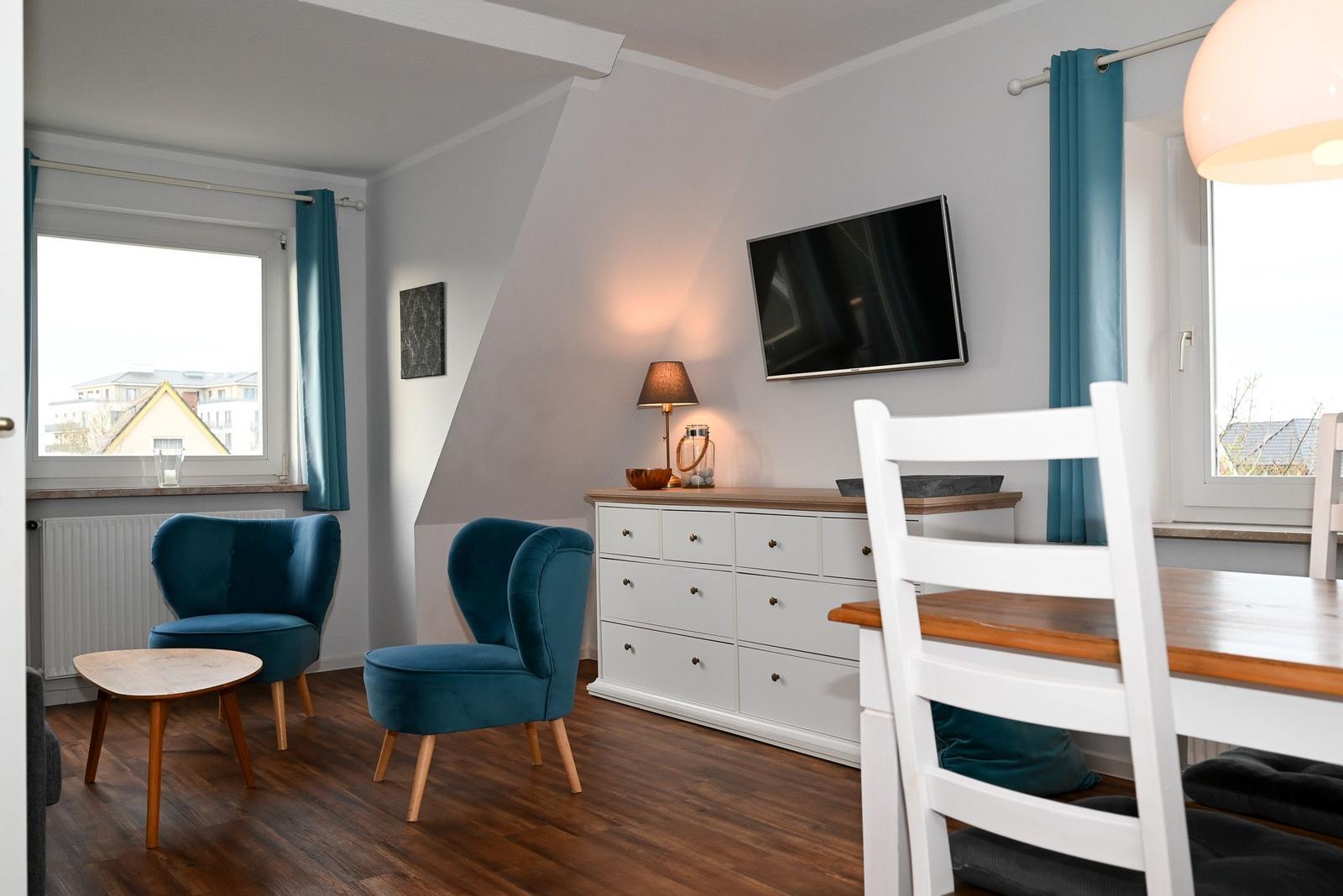 Zimmer mit zwei blauen Sesseln, Schreibtisch, TV und Holzboden.