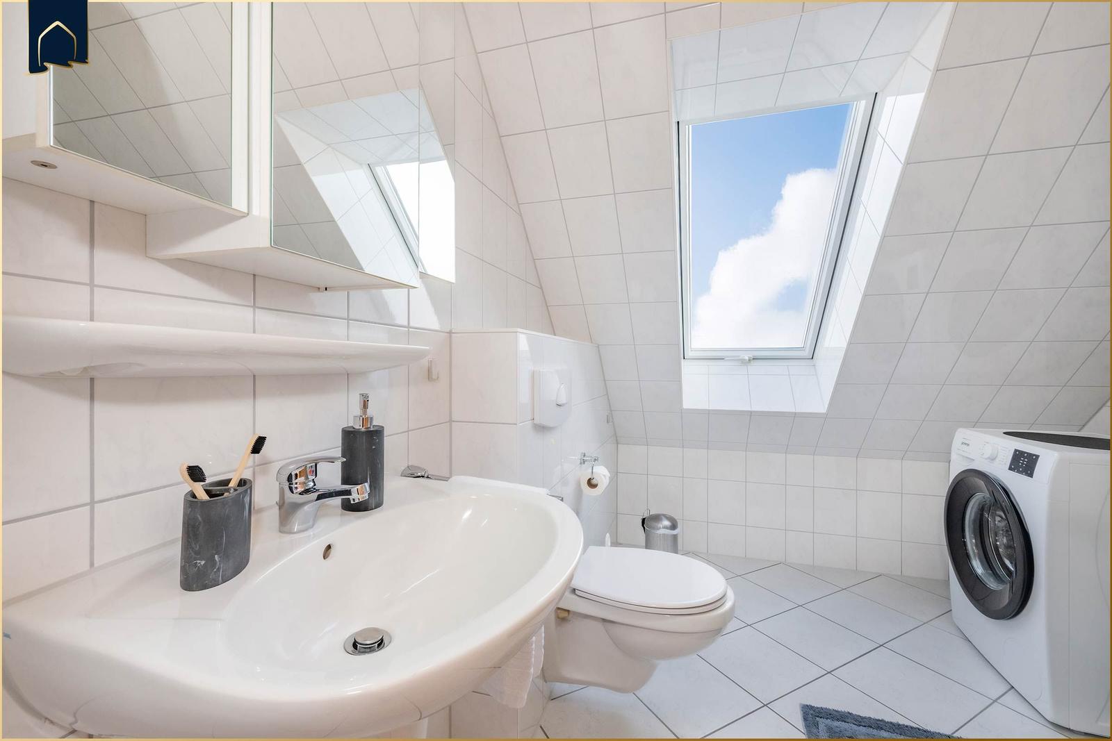 Badezimmer mit Waschmaschine, Toilette und Dachfenster. Weiße Fliesen und Spiegel.