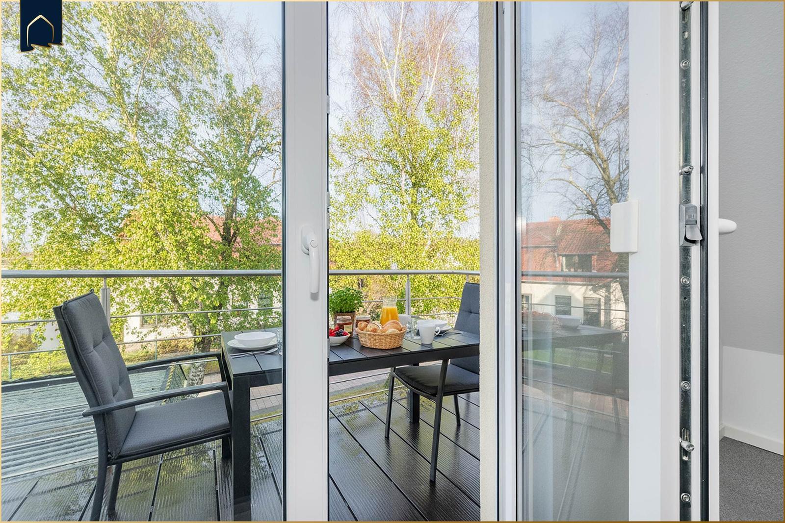Terrasse mit Tisch, Stühlen und Frühstücksgeschirr. Blick auf Bäume und Gebäude.