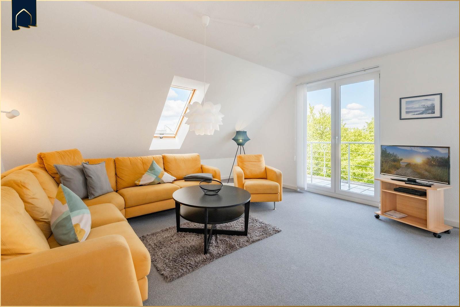 Wohnzimmer mit gelbem Sofa, Rundtisch und TV. Balkon und Dachfenster mit Blick.