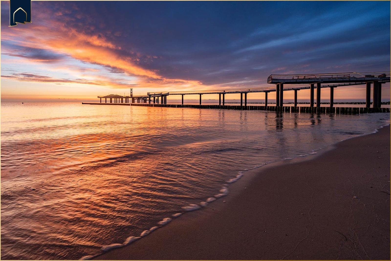 Sonnenuntergang am Strand mit Blick auf einen langen Steg ins Wasser.
