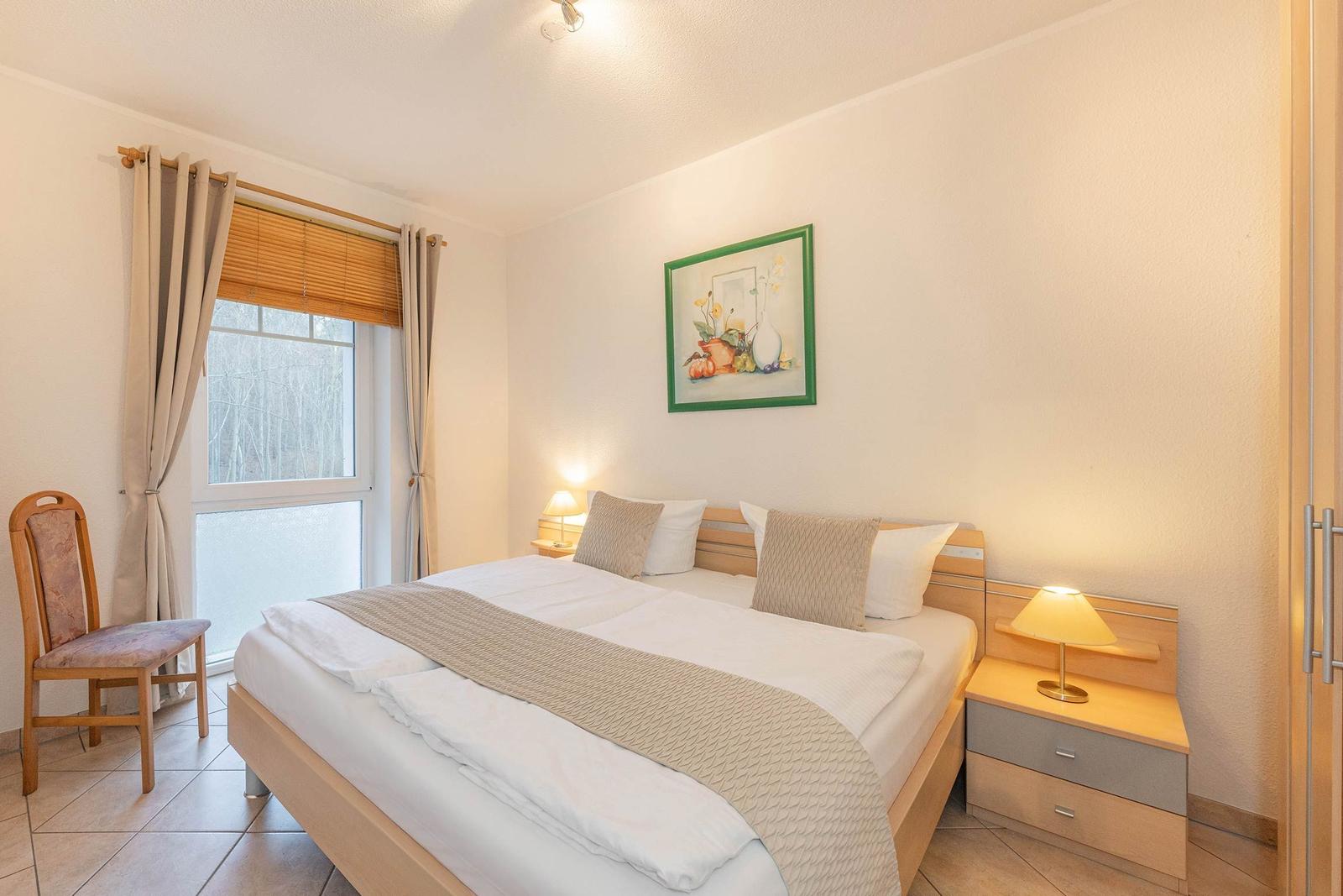 Schlafzimmer mit Doppelbett, Nachttisch, Stuhl und Fenster mit Blick auf Wald.
