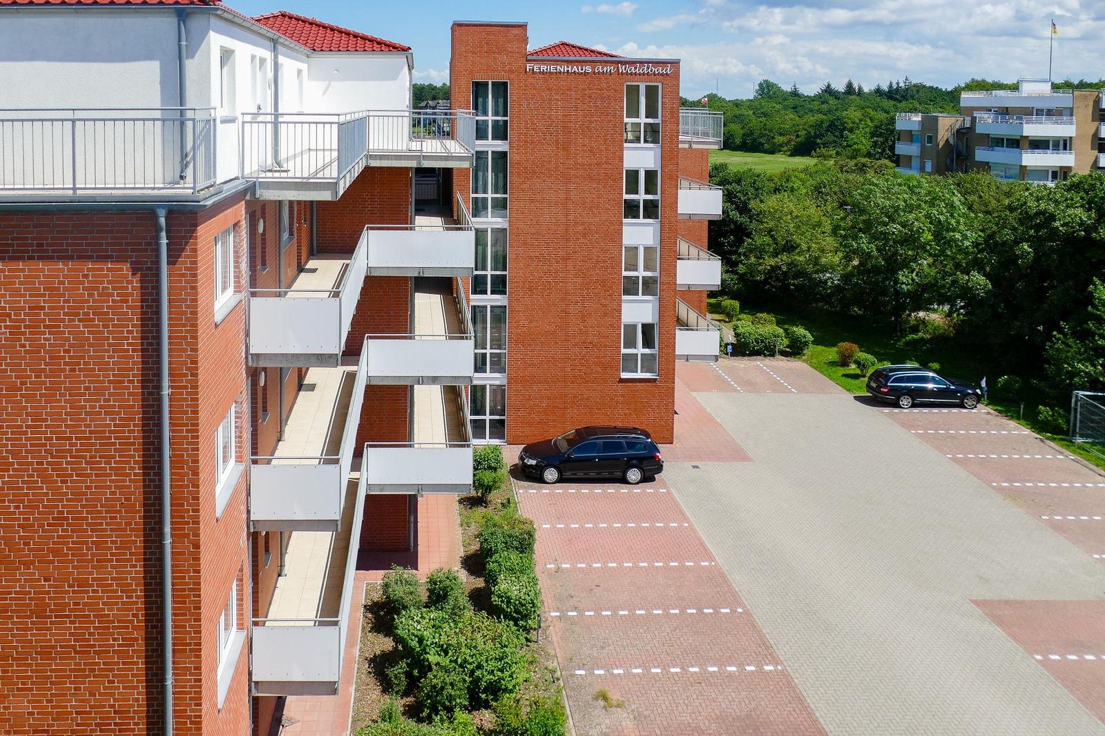 Ferienhaus am Waldbad mit Balkonen und Parkplatz. Grünanlagen und Wald im Hintergrund.