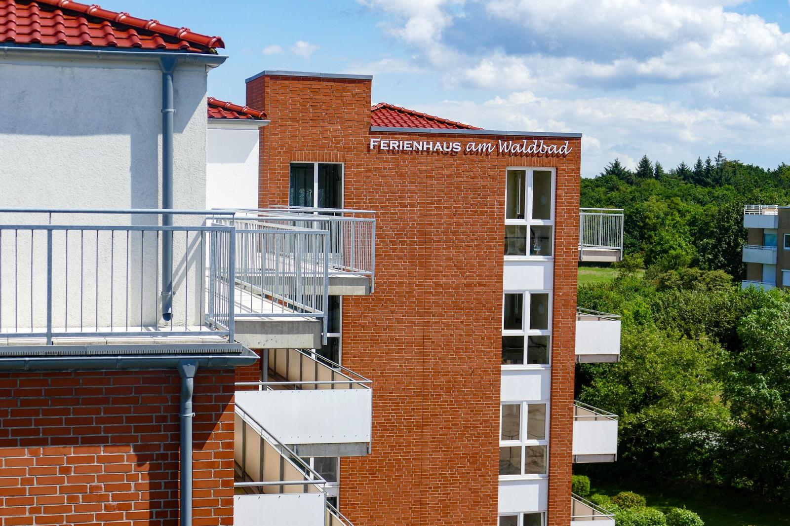 Ferienhaus am Waldbad mit Balkonen und rotem Dach unter blauem Himmel.