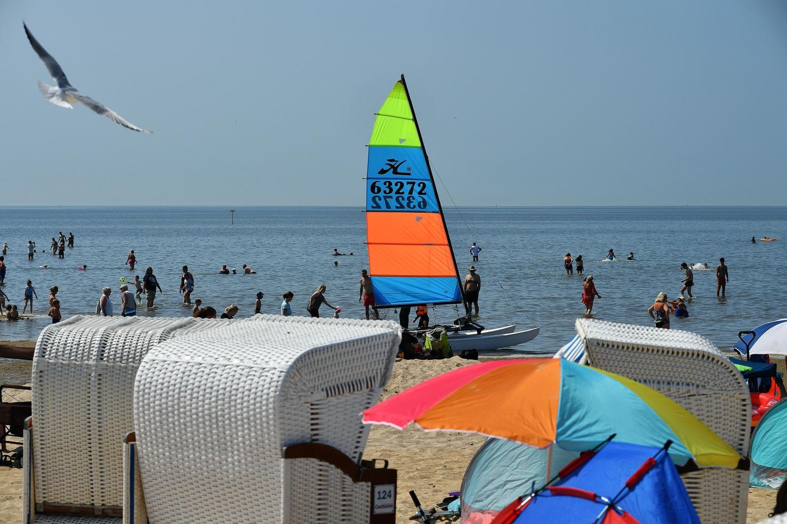 Strand mit weißen Liegestühlen, buntem Sonnenschirm und Segelboot im Wasser.