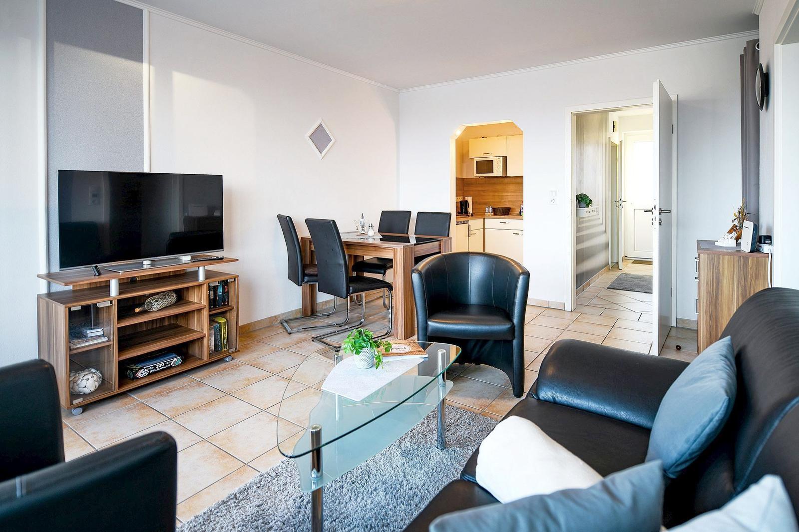 Wohnzimmer mit Sofa, Sessel, Esstisch und TV. Küche im Hintergrund sichtbar.