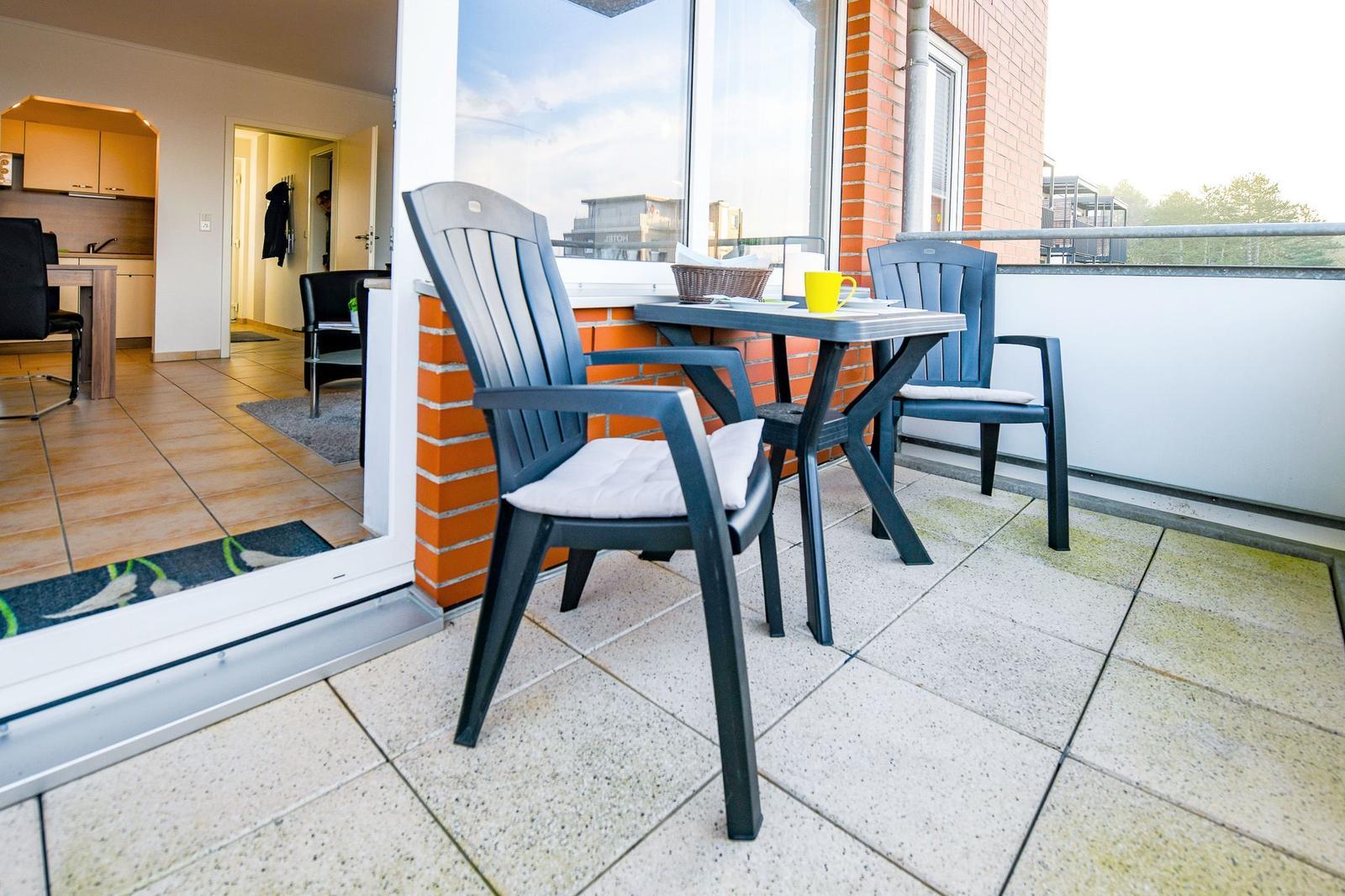 Terrasse mit Tisch und zwei Stühlen. Blick ins Wohnzimmer mit Küche.