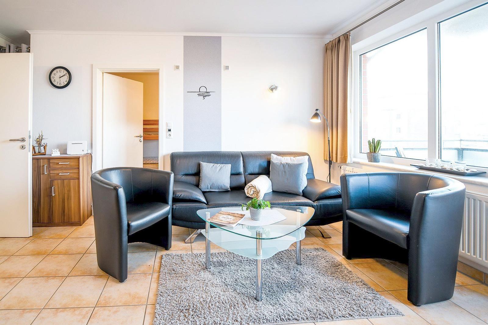 Wohnzimmer mit Ledersofa, zwei Sesseln und Glas-Tisch. Fenster mit Vorhängen und Teppich.