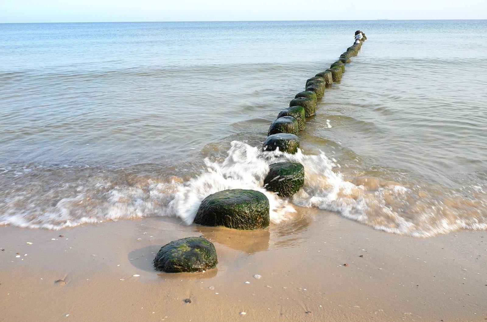 Wellen brechen an einer Reihe von grünen Steinen am Strand.