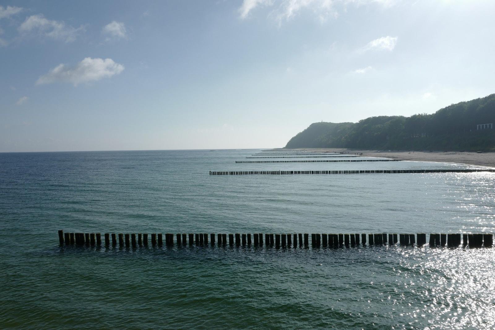 Meer mit Holzpfählen und grüner Küstenlinie unter blauem Himmel.