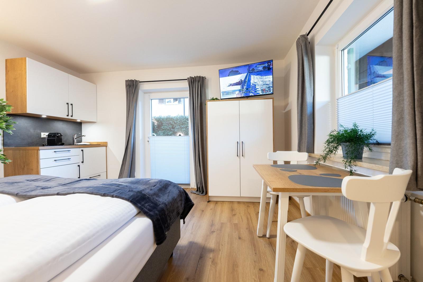 Schlafzimmer mit Küchenzeile, Esstisch und Fernseher. Holzboden und große Fenster.