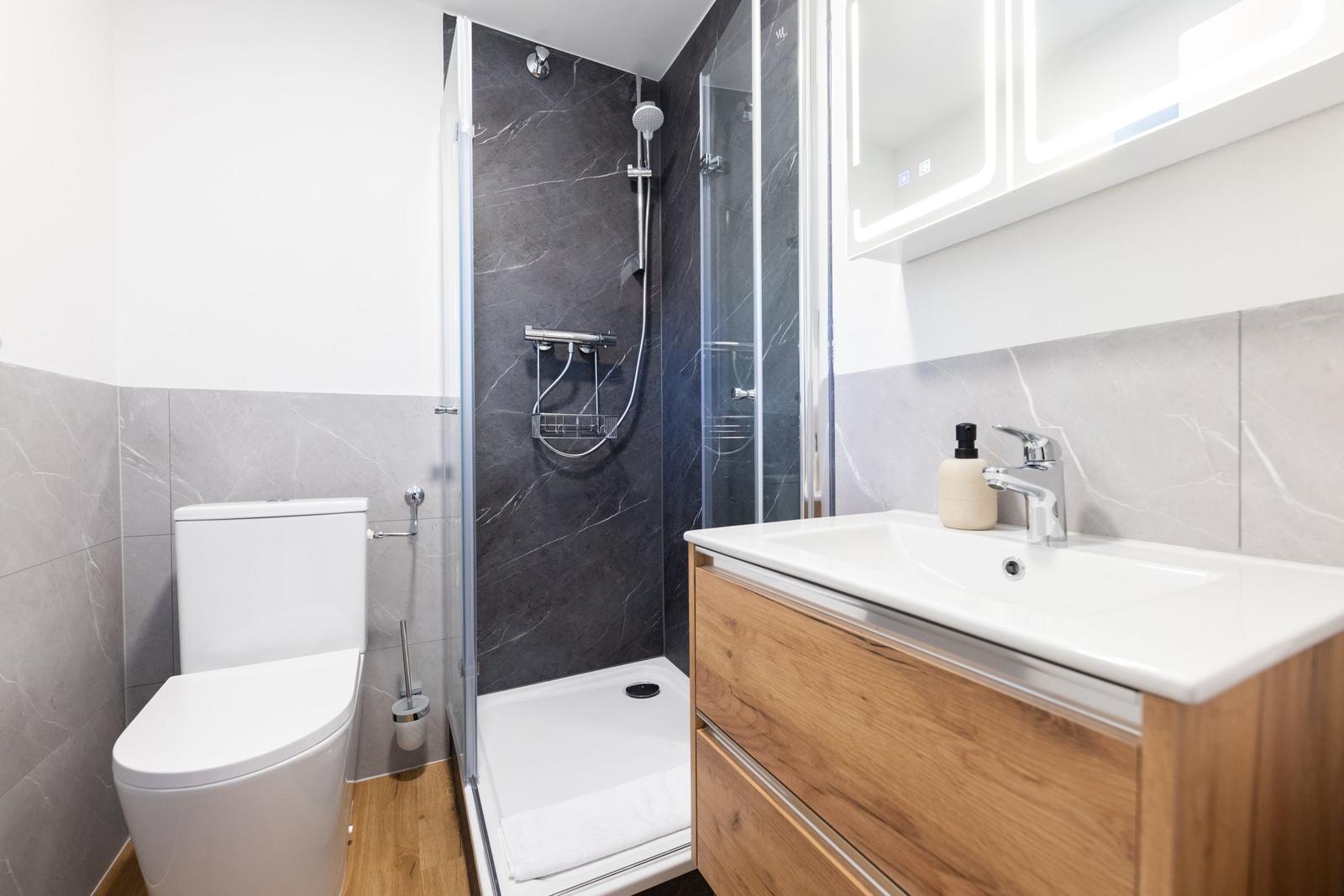 Badezimmer mit Dusche, Waschbecken und Toilette. Marmorfliesen und Holzschrank.