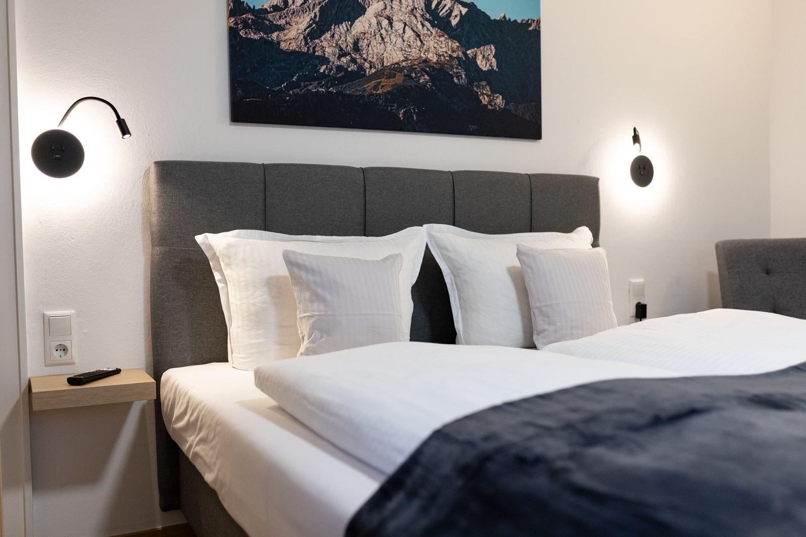 Schlafzimmer mit Bett, Kopfteil und Wandbild. Beleuchtung und Nachttisch sichtbar.