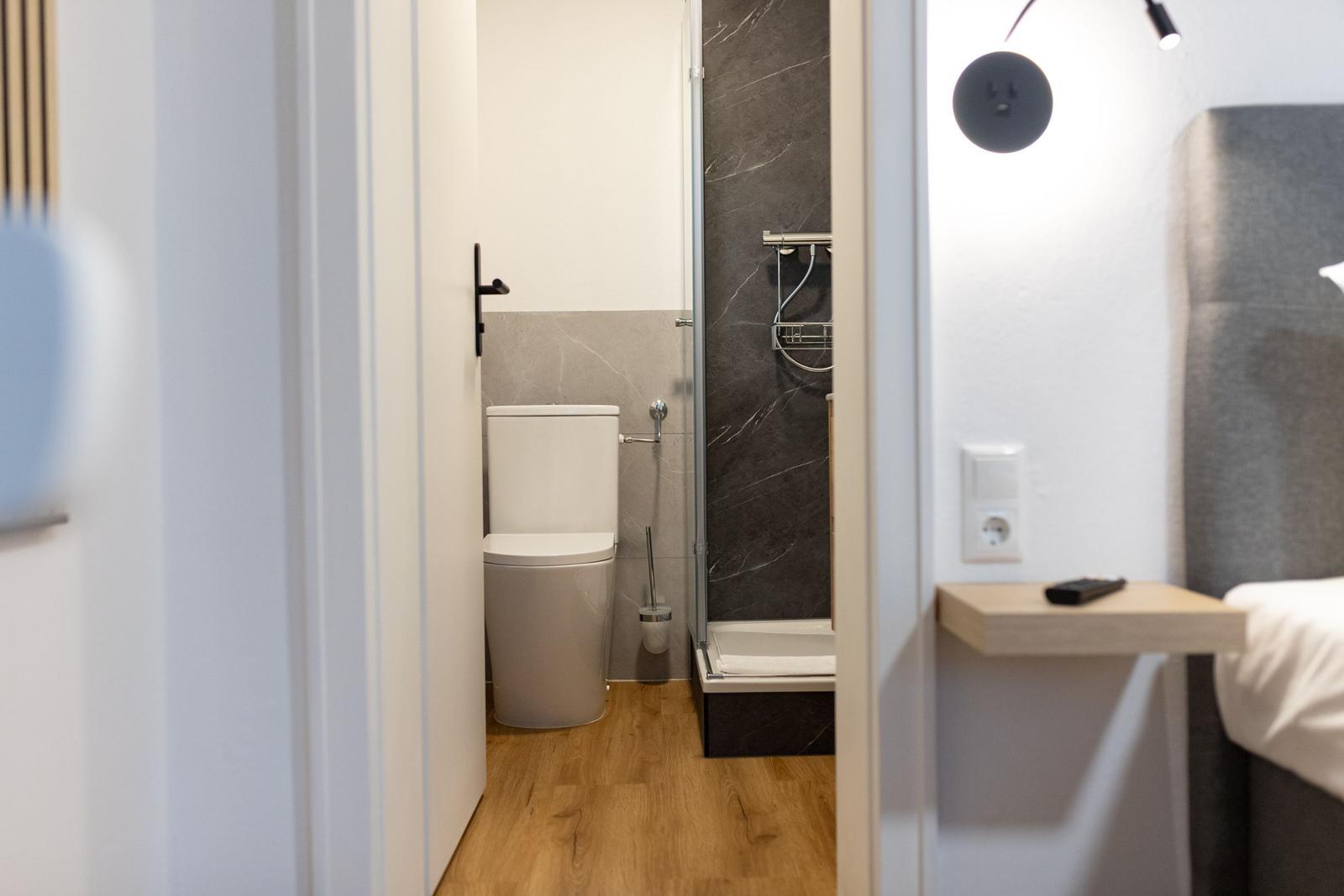 Badezimmer mit Dusche und Toilette, Holzboden und Nachttisch neben dem Bett.